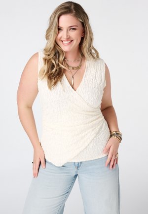 Lachende vrouw met golvend blond haar die een mouwloze witte gestructureerde top, lichtblauwe spijkerbroek, gelaagde kettingen en armbanden draagt.