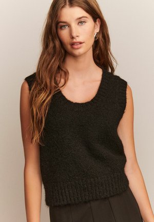 Jongeman met lang bruin haar, gekleed in een mouwloos zwart gebreid sweater, gecombineerd met zwarte broeken tegen een effen achtergrond.