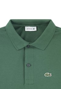 Zielona koszulka polo z teksturowanej tkaniny, z płaskim kołnierzykiem, dwoma guziczkami oraz małym, haftowanym logo krokodyla na lewej piersi.