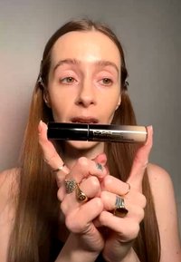 Frau mit langen Haaren hält den MAC Studio Fix Concealer und zeigt das Produkt nah an der Kamera mit mehreren Ringen an den Fingern.