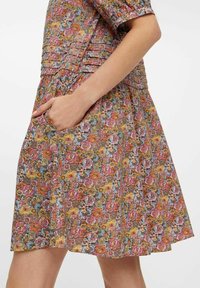 Sukienka w kwiatowy wzór w wielu kolorach; z plisowanym fasonem, krótkimi rękawami i bocznymi kieszeniami. Wykonana z lekkiego materiału o gładkiej teksturze.