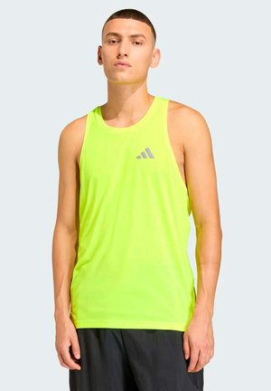 Jonge man met kort haar, gekleed in een neongeel Adidas mouwloos shirt en zwarte korte broek, staand tegen een effen grijze achtergrond.