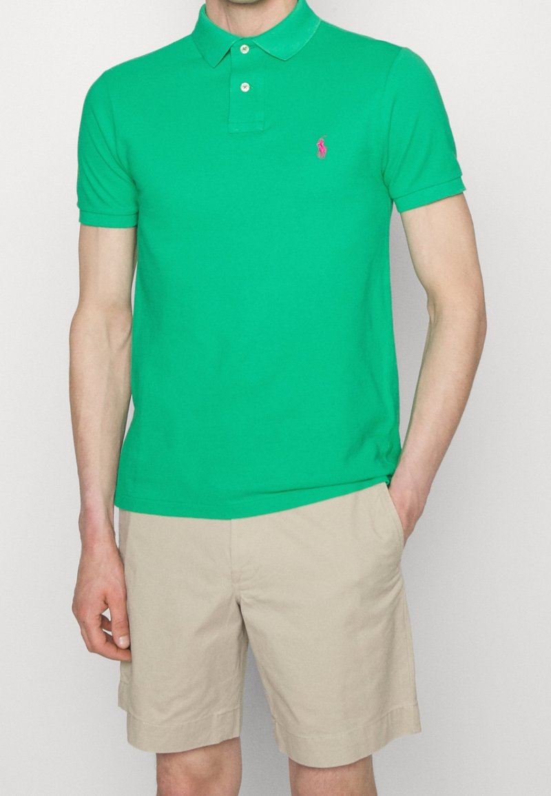 Polo en coton vert à manches courtes, patte de boutonnage à deux boutons et petit logo rose sur la poitrine gauche. Porté avec un short beige.