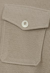 Beiges, geripptes Gewebe mit kleinem Taschenklappe und einem weißen Knopfdetail. Die Textur ist weich und das Design schlicht und funktional.