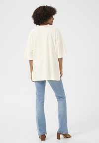 Lös passform på en krämfärgad t-shirt med korta ärmar, kombinerad med ljusblå utsvängda jeans och bruna klacksandaler, sedd bakifrån.