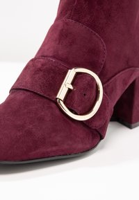 Burgundy mockaskäl med fyrkantig tå, guldfärgad metallaccenter och lagerdesign på remmen. Blockklack och slät sula.