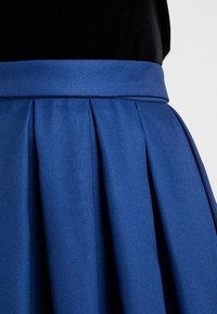 Sininen laskostettu hame, joka on valmistettu tekstuuriin omaavasta kankaasta ja jossa on sileä vyötärönauha. Kankaan pinta heijastaa hienovaraista kiiltoa, joka syventää väriä.