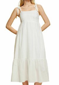 Femme portant une robe midi blanche sans manches avec un corsage brodé, une jupe à volants et des bretelles nouées aux épaules, debout les mains derrière le dos.