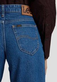 Vue arrière d'un jean Lee bleu avec une étiquette de marque en cuir et un détail de couture courbée sur la poche, associé à une manche de chemise marron foncé avec broderie.
