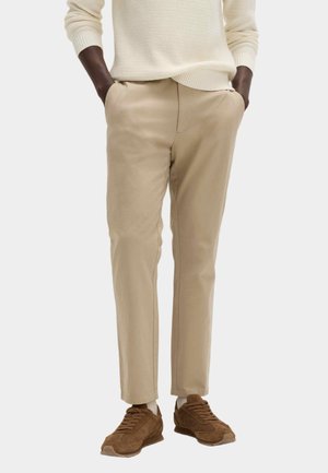 Person iført beige bukser med lige pasform, lysebeige striktrøje og brune ruskindssneakers, hænderne i lommerne, stående mod en ensfarvet baggrund.