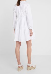 Bílá bavlněná šaty s dlouhými rukávy, na rameni řasená, s vrstveným spodním lemem a volným střihem. V kombinaci s bílými espadrilkovými sandály na klínu.