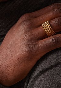 Goudkleurige ring met een gestructureerd, rechthoekig patroon, gedragen op een hand met een donkere huid die op een grijze stoffen ondergrond rust.