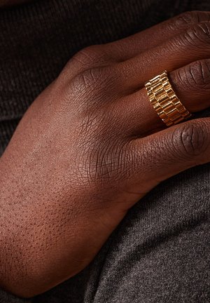 Goudkleurige ring met een gestructureerd, rechthoekig patroon, gedragen op een hand met een donkere huid die op een grijze stoffen ondergrond rust.