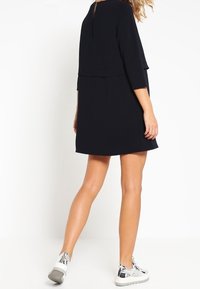 Abito blu navy con maniche a tre quarti e design a strati. Indossato con sneakers argentate, che mostrano una texture liscia e una silhouette semplice.