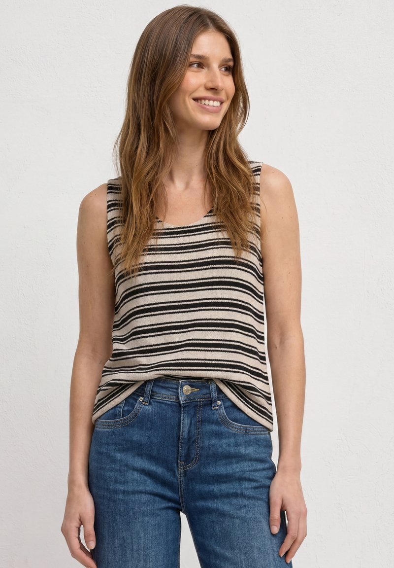 Jeune femme souriante, portant un haut beige sans manches avec des rayures noires et un jean bleu taille haute, debout devant un fond blanc uni.