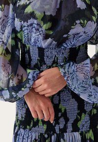 Bloemenjurk met doorschijnende pofmouwen, een smocktailie in groene en blauwe tinten, met een vloeiende rok en gedetailleerde bloemenpatronen.