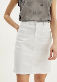 Kvinde iført en hvid denimnederdel med forlommer og bæltestropper, parret med en grå udenærmet top med små blomsterapplikationer.