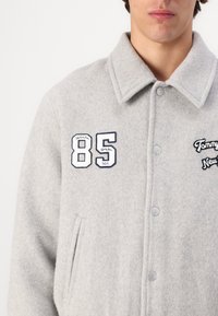 Giacca in misto lana grigia con colletto, caratterizzata da accenti ricamati "85" e "Tommy Jeans" sul petto. Chiusura anteriore con bottoni.