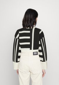 Pull court avec de larges rayures géométriques noires et crème, doté de manches larges. Porté avec un pantalon en velours côtelé crème et une étiquette de marque.