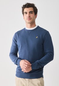 Mann iført en marineblå sweatshirt med lite gult fuglelogo, lagd over en hvit skjorte, som står foran en enkel lys bakgrunn.