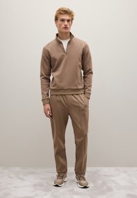 Brauner Reißverschluss-Hoodie mit gerippten Bündchen, kombiniert mit passenden, taillierten Hosen. Das Outfit wird mit neutrofarbigen Sneakern gestylt. Glatte Stofftextur.