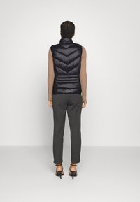 Schwarze Pufferweste mit hohem Kragen und Zickzacksteppung, getragen über einem beigen ribbelten Pullover, kombiniert mit grauen Hosen und Riemchenschuhen.