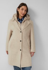 Beige jas met een capuchon, gestructureerd ontwerp, sluiting met één knop, zijzakken en een textuurstof. Draagt over een geruite blouse.