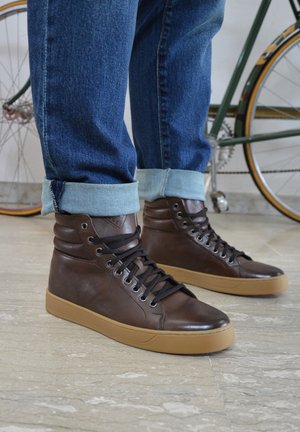 Bruine high-top sneakers met een glad leren bovenwerk, zwarte veters en een tan rubberen zool. De spijkerbroek is opgerold bij de enkels, waardoor de schoenen duidelijk zichtbaar zijn.