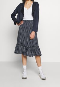 Giacca corta in suede blu navy con fronte a cascata. Top bianco con scollatura a V, gonna A-line a pois con orlo arricciato e sneakers bianche.