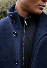 Cappotto di lana blu navy con colletto alto, dotato di bottoni neri e una giacca nera a strati con zip sottostante, che mostra una texture elegante.