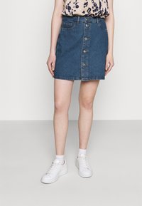 Denim skirt with a straight cut, front button closure, and five pockets. Worn with a patterned top and white sneakers.
