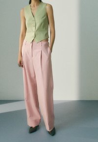 Jasnozielony bezrękawnik z guzikami w połączeniu z różowymi szerokimi spodniami. Tekstura wydaje się gładka, spodnie mają boczne kieszenie.