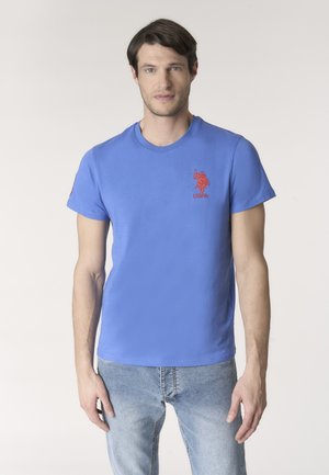 Uomo che indossa una maglietta blu USPA con logo rosso sul petto e jeans blu sbiaditi, in piedi contro uno sfondo chiaro e uniforme.