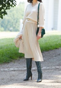 Femme debout à l'extérieur, portant une jupe plissée beige, un pull crème, des bottes noires jusqu'aux genoux, tenant un manteau beige et portant un sac à épaule vert.