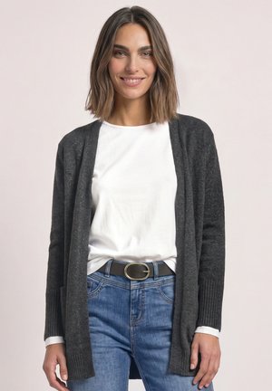 Femme aux cheveux bruns mi-longs sourit, portant un cardigan gris foncé, une chemise blanche, un jean bleu et une ceinture noire avec une boucle ovale dorée.