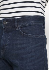 Tmavě modré džíny z denimu s knoflíkovým zapínáním, designem s pěti kapsami a světlým prošíváním. Doplňují se pruhovaným topem v námořnické modři a bílé.