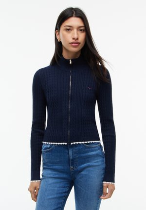 Жена, облечена в тъмносин свитър с цип и релефен десен с изрези по краищата и сини дънки на едноцветен фон.