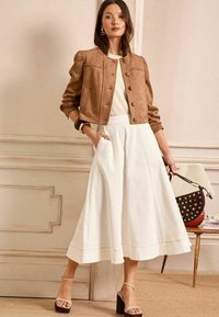 Suède bruine cropped jack met knoopsluiting, gecombineerd met een witte midi-rok met zijzakken. Model houdt een ronde handtas vast.
