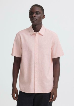 Homme portant une chemise à manches courtes rose clair avec des boutons et un pantalon noir, debout devant un fond blanc uni.