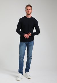 Granatowy sweter z długim rękawem o teksturowanym wzorze, zestawiony z jasnoniebieskimi dżinsami i białymi sneakersami. Model stoi z rękami z przodu.