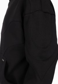 Schwarzer Stoff mit gepolsterten, gesteppten Ellbogenpatch-Details auf einem Langarmkleidungsstück, das Textur und Nahtlinien zeigt.