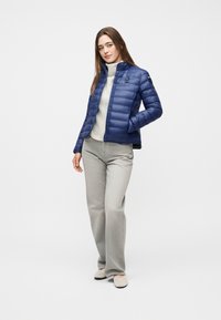 Γυναίκα που φοράει μπλε μπουφάν puffer πάνω από λευκό πουλόβερ ζιβάγκο, γκρι παντελόνι και μπεζ παπούτσια, στέκεται με το ένα χέρι στην τσέπη της.