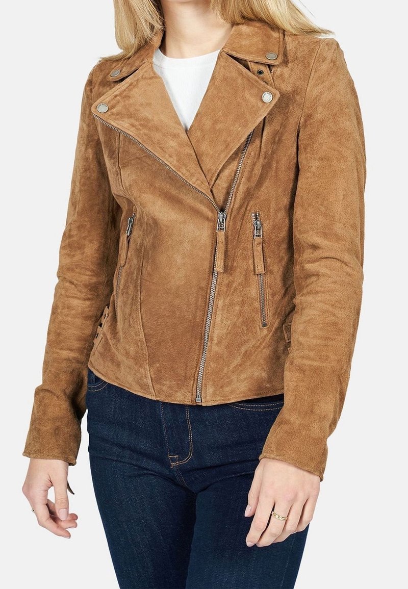 Femme portant une veste de motard en suède beige avec une fermeture éclair asymétrique à l'avant et des poches zippées, associée à un jean bleu foncé.