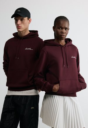Deux modèles portant des sweat-shirts à capuche bordeaux foncé assortis avec poches avant ; la femme porte une jupe plissée blanche, l'homme porte un pantalon noir et une casquette noire.