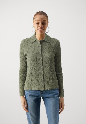 Texturovaný olivově zelený cardigan s dlouhými rukávy, zapínáním na knoflíky a límcem. Spárováno s modrými džíny.