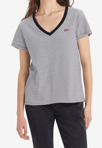 Kobieta w czarno-białą stylizowaną koszulkę z dekoltem w serek i krótkimi rękawami, w czarnych spodniach, stojąca z rękami swobodnie wzdłuż ciała.