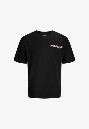 Usmievajúci sa chlapec stojaci, oblečený v čiernom tričku s potlačou, svetlomodrých džínsoch a čiernych topánkach na bielom pozadí.