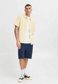 Korten mouwen beige overhemd met knopen over een witte T-shirt, gecombineerd met marineblauwe shorts. Model draagt zwarte sneakers met crèmekleurige zolen en witte sokken.