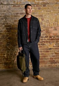 Dark denim jacket with front pockets, paired with a red sweatshirt and wide-leg jeans. Brown boots and an olive green bag complete the look.