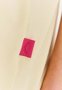 Béžová látka s hladkým povrchem, která má výraznou růžovou etiketu s stylizovaným designem řasy přišitou do švu.